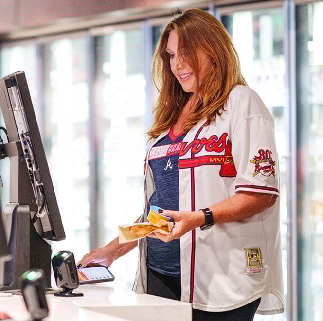 A woman wearing a baseball jersey purchases snacks inside a stadium using her cell phone to pay.