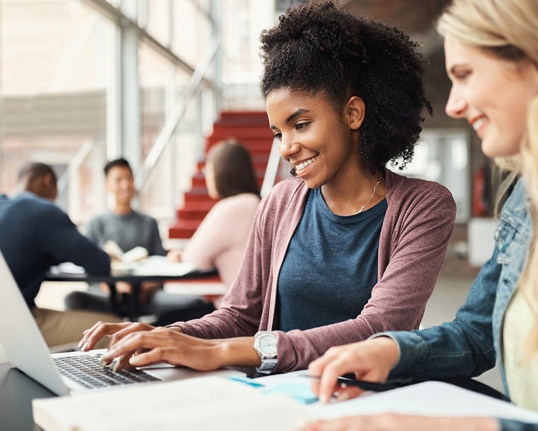 Two young women sit at a table in a common area of a university building and look at a laptop screen together.