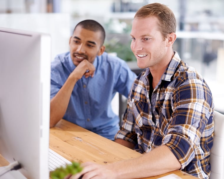 Two young men in button-down shirts sit at a desk and smile as they look at a computer screen together.