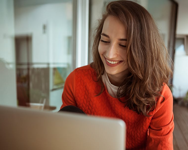 A young woman in a bright sweater smiles as she looks at a laptop screen.