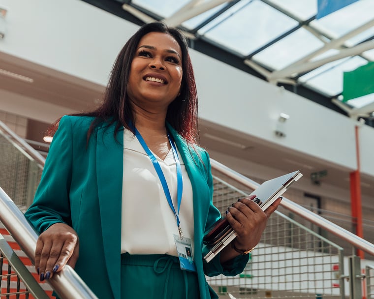 A woman wearing a green blazer and blue lanyard smiles as she walks down a staircase in a large building.