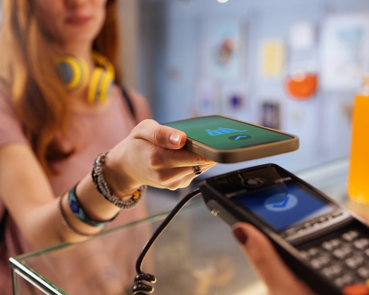 A teenager wearing headphones taps her cell phone on a checkout terminal.