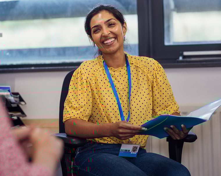 A smiling woman in a yellow, polka dot shirt is sitting in a chair and holding a folder.