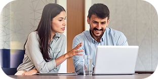 A man and a woman sit and look at a laptop screen together in an office setting.