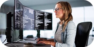 A woman in an office looks at several large computer monitors.