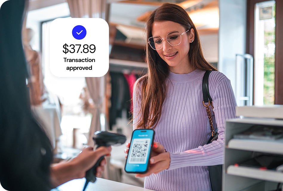 A woman in a store holds her cell phone, displaying a QR code. The cashier scans the QR code to complete the transaction.