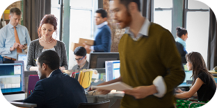A busy workplace shows several people sitting at computers or moving around the office carrying stacks of paper or using smartphones. 