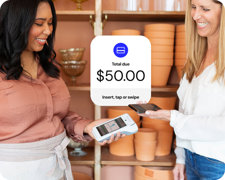 Two women smile in a flower shop as one makes a payment via smartphone to a handheld device. 