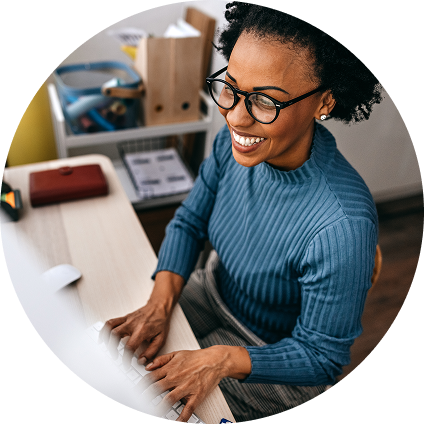 A smiling woman wearing a blue sweater and glasses works at her desk.