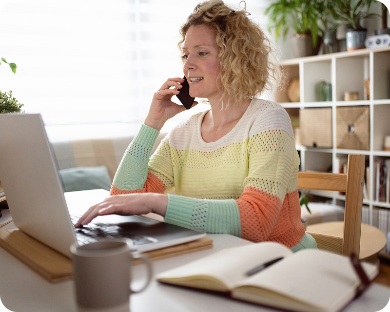 A woman in a colorful sweater sits at a desk using a laptop while talking on a phone.