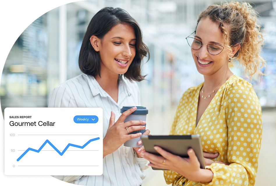 Two businesswomen look at a sales report in an office setting. One of them is holding a coffee, the other is holding a tablet.