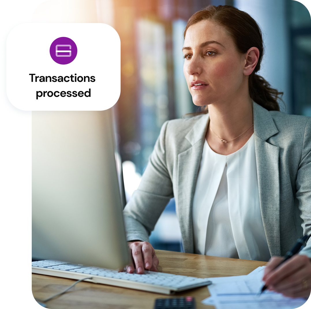 A woman sits at a table with a desktop computer and a graphic overlaying says transaction processed.