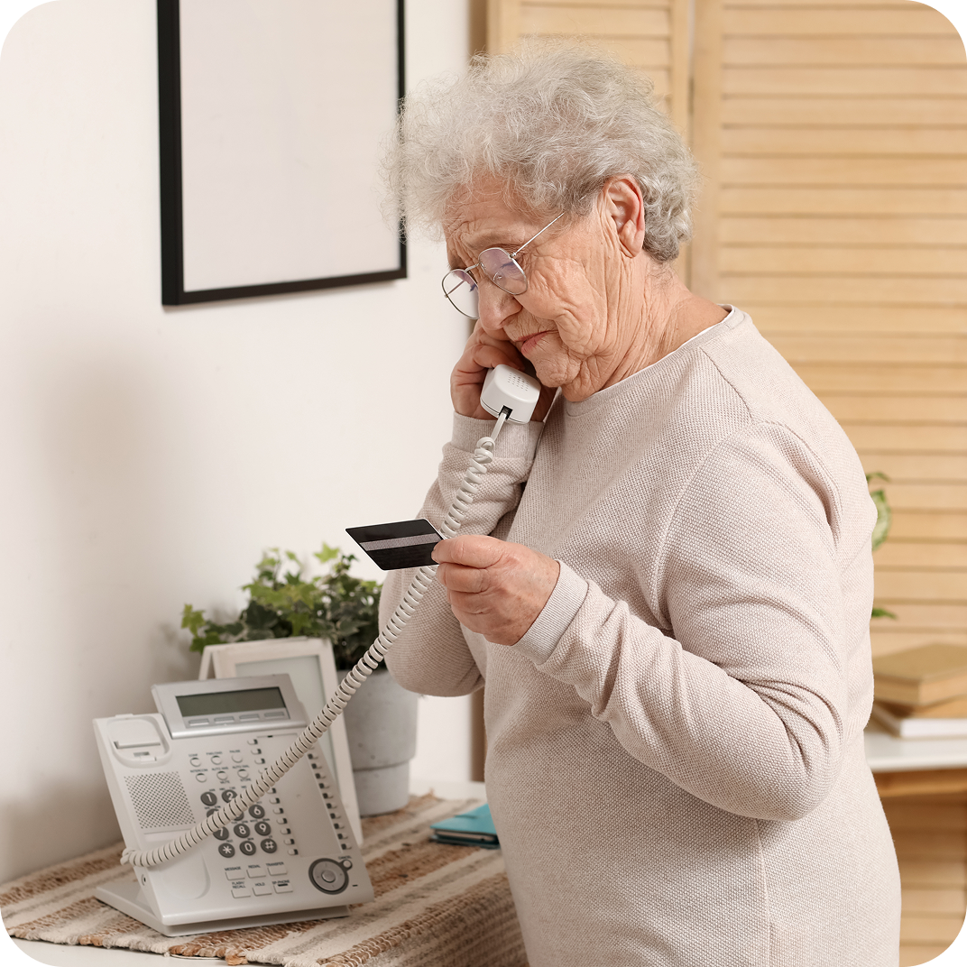 A standing woman speaks over a landline phone to make an IVR payment with the credit card in her hand.