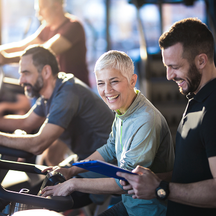 A group of people participating in a fitness class. A woman is talking with a trainer holding a clipboard.