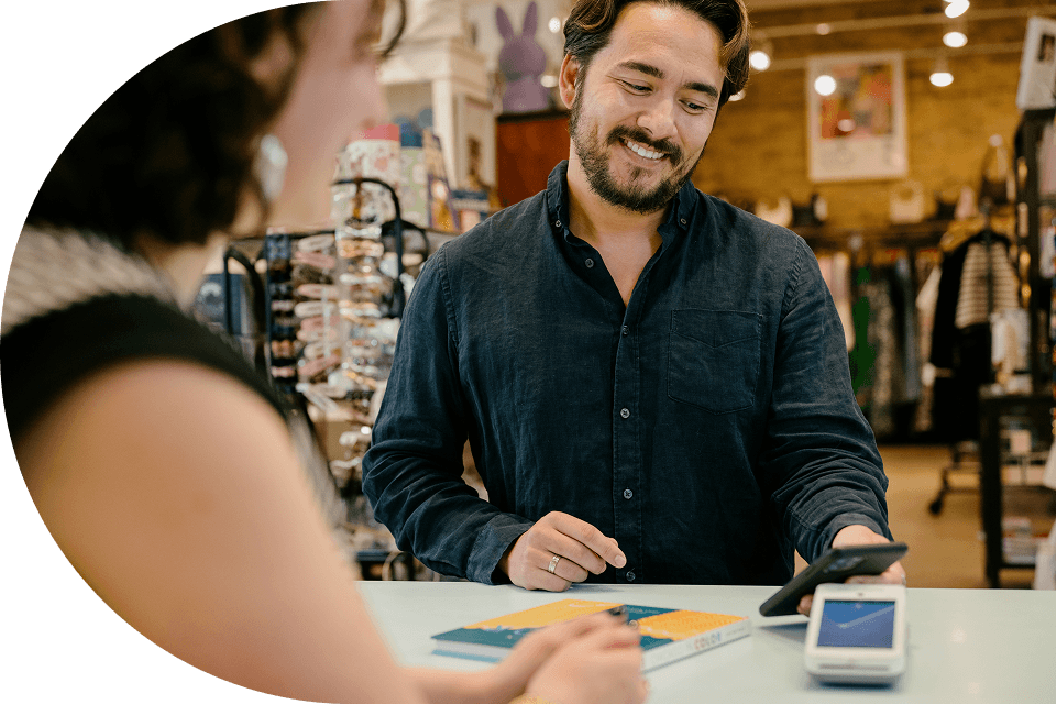 An offscreen customer taps their card to pay for a purchase on the customer-facing side of a Genius countertop POS. A small business employee stands on the other side of the counter tapping the employee-facing screen of the POS.