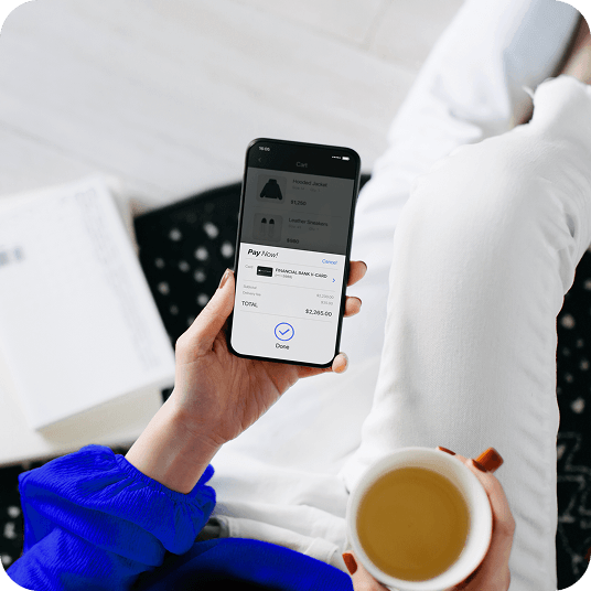 Overhead shot of a person completing an online transaction on their phone using Apple Pay. They are sitting on a couch and holding a cup of coffee