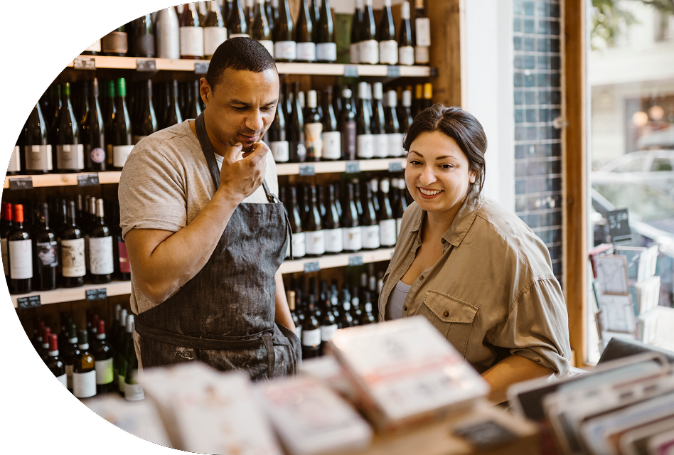 A staff member in an apron helps a customer who is browsing items at a wine store.