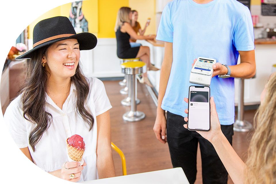 A customer at an ice cream shop eats an ice cream cone while her friend pays for it on a card machine from a phone mobile.