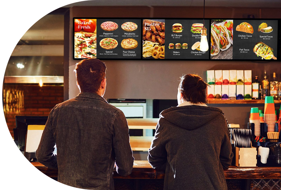 Two people stand at a counter of a quick service restaurant with their backs toward the camera as they look at the vibrant offering on an overhead indoor digital menu board.
