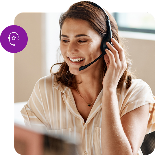 A smiling customer service representative wearing a headset assists someone over the phone while sitting at her computer.