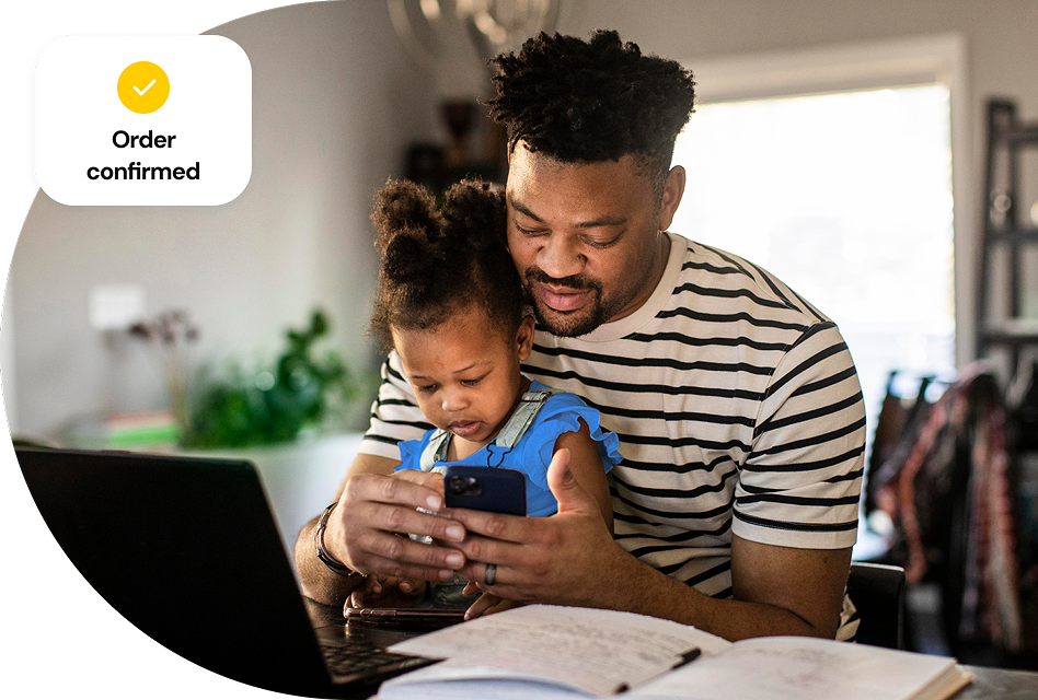 A man sitting at his home in front of an open laptop holds his daughter in his lap as he looks at a phone, while a small graphic overlaying the image shows an order confirmed graphic.