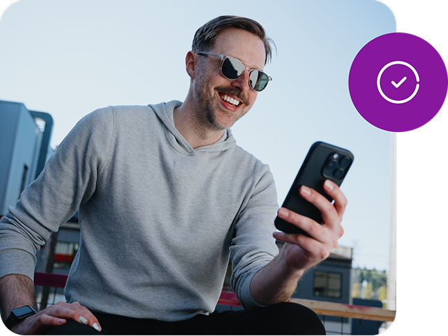 A smiling man in sunglasses looks down at a phone in his hand and a small, round, graphic icon showing a checkmark overlays the image.