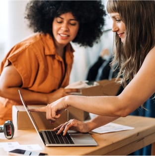 Two business women pack boxes and scruitinise inventory on a laptop.