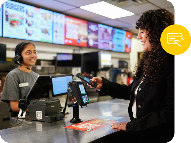 A digital menu board lights up the background behind a fast food worker, accepting a point of sale payment.