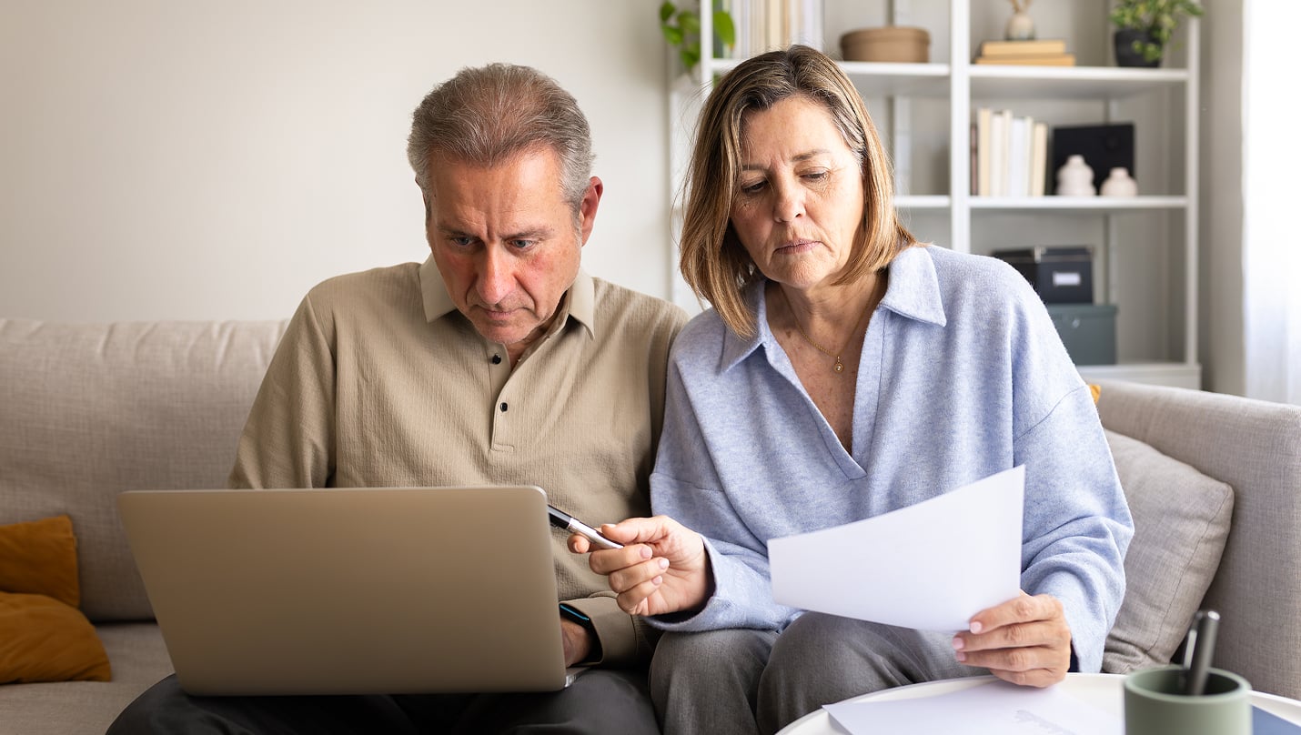 Two people sitting together on a couch reviewing bills on a laptop and paper statements.