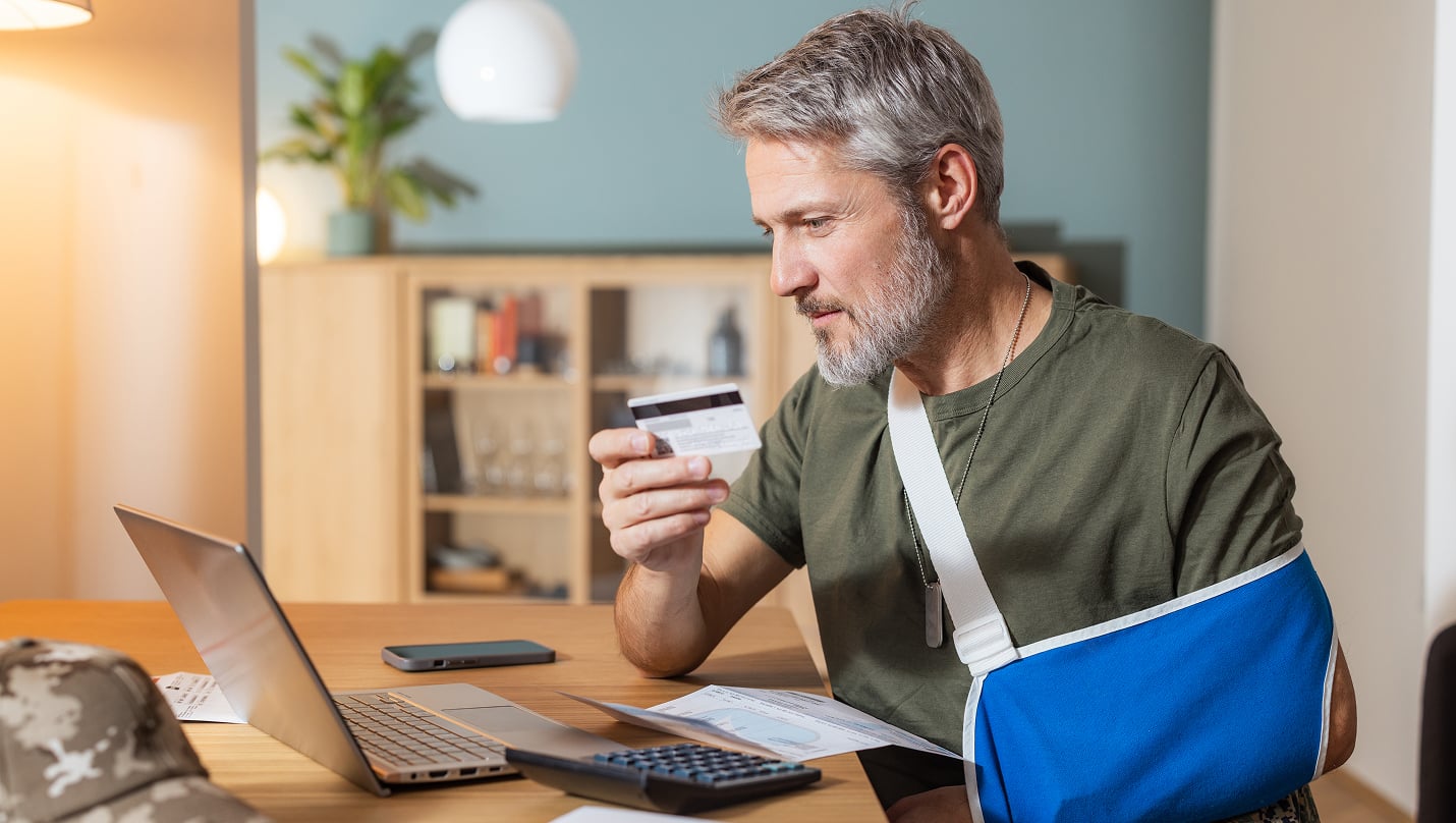 A person with an arm in a sling using a laptop while holding a credit card at a table.