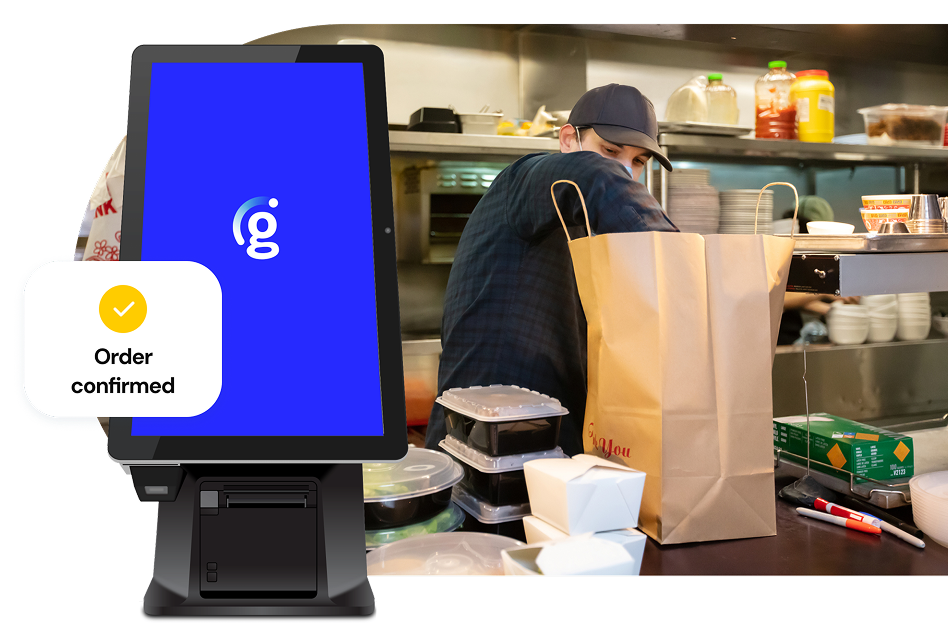 A look at a busy kitchen scene featuring a person stacking takeaway containers in a large brown bag; a Genius kiosk with a small UI call-out that says “Order confirmed” sits in the foreground.