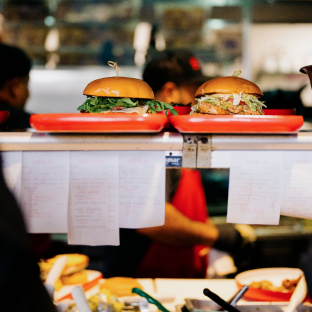 Two burgers sit on red trays atop a kitchen ticket holder, while an employee in a red apron works in the background. 