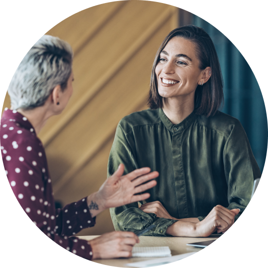 Two employees sit at a desk, one is smiling and listening and one is explaining something.