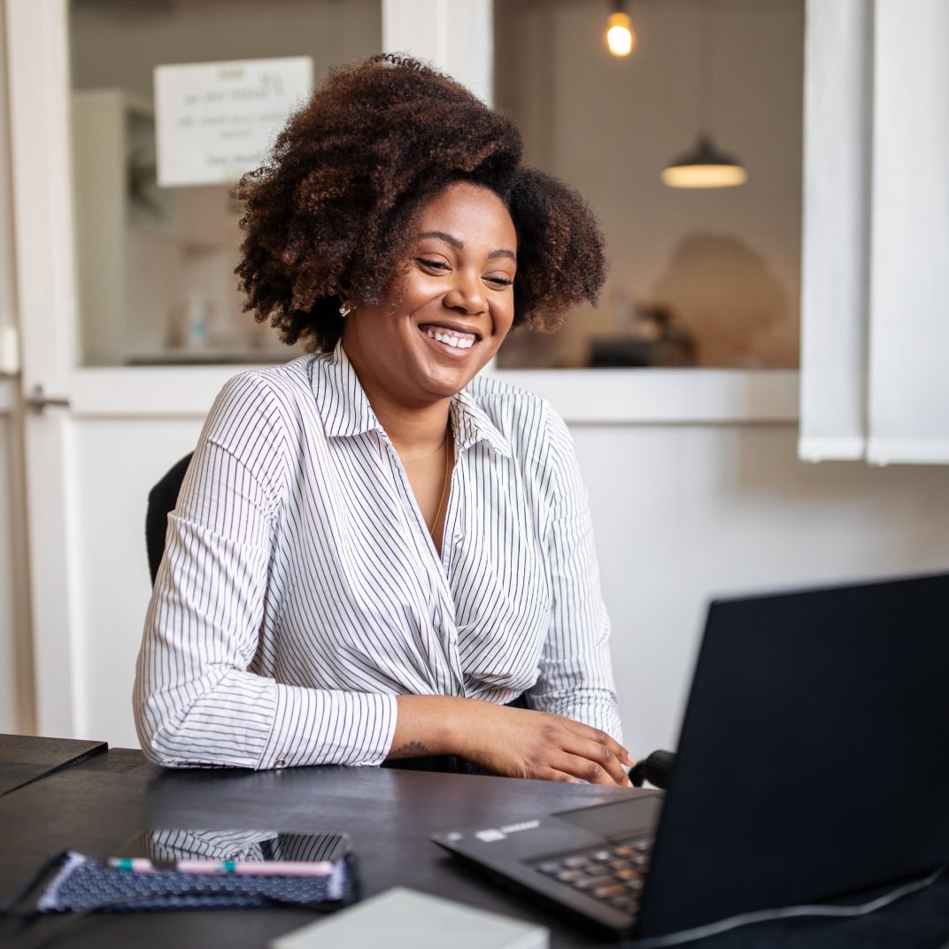 A smiling woman works at a desk, with her laptop opened in front of her.