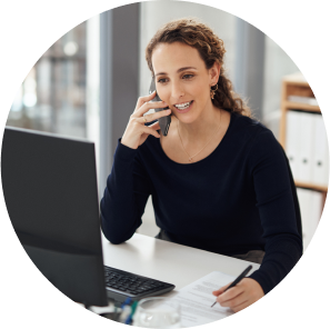 A woman sitting in a corporate office in front of her computer monitor talks on the phone and takes notes with her left hand.