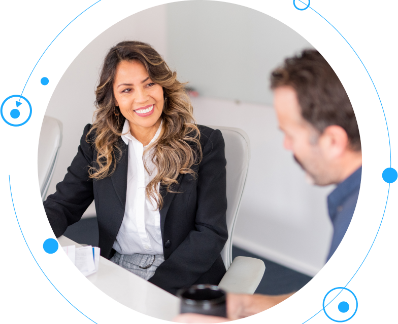 A smiling woman wearing a blazer talks to her colleague in a meeting room.