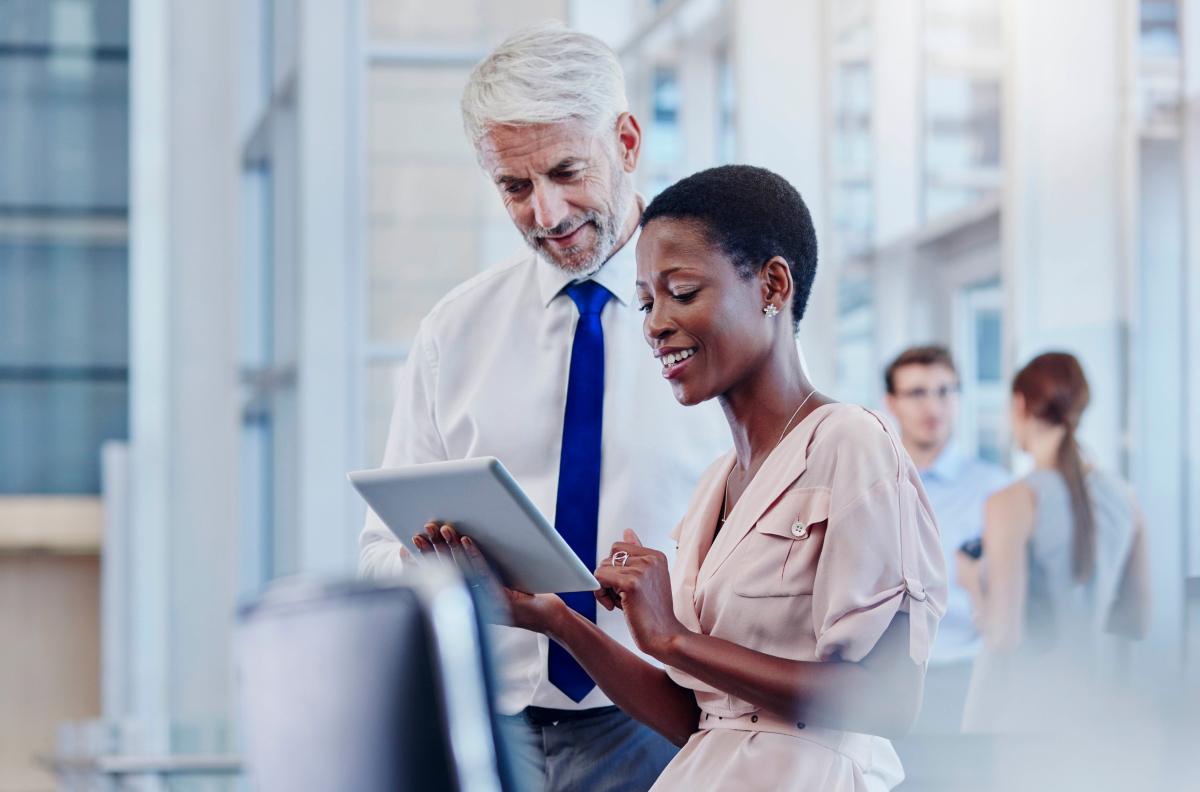 A man and woman stand in a busy office looking at a large tablet.
