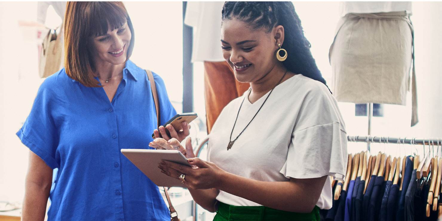 Smiling customer holds smartphone while looking into tablet device held by smiling retailer