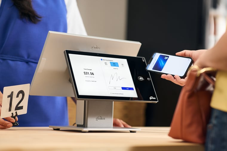 Close-up of hand holding smartphone to tap to pay at countertop point of sale device as merchant looks on