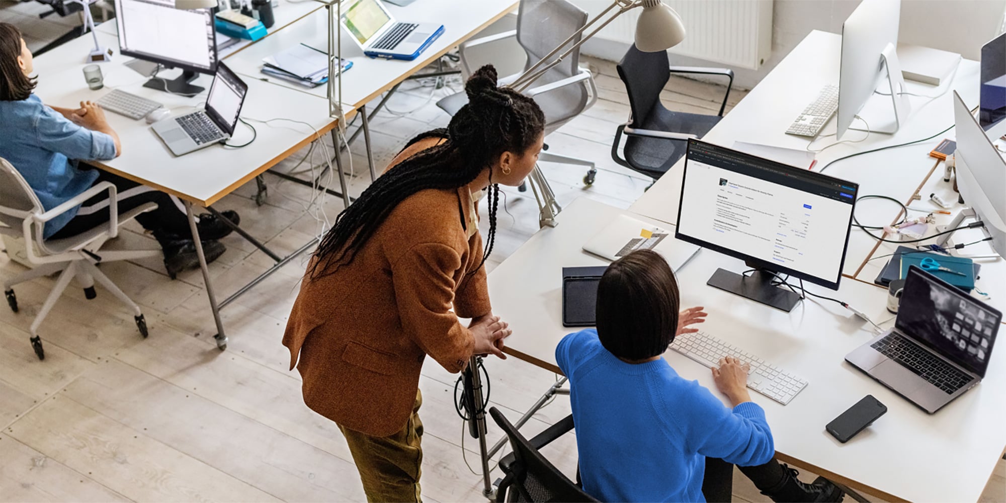 Person in brown blazer standing near seated colleague in blue sweater while collaborating at desk in modern office
