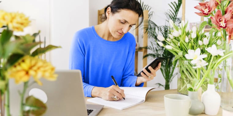 Seated person using pen to write in notebook while holding phone device