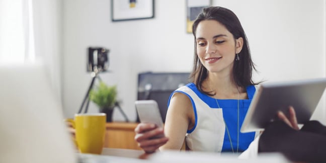 Woman sitting at work desk smiling holding tablet in one hand and mobile phone in the other