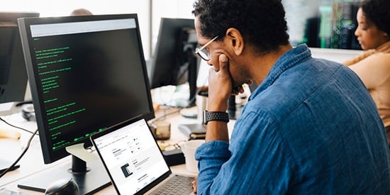 A man sitting at a desk looks at a laptop computer in front of a larger computer monitor.