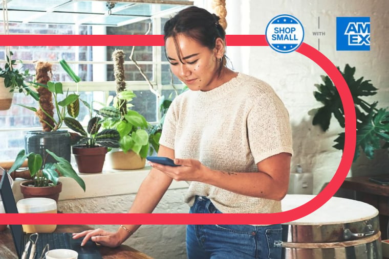Retail worker stands at desk with one hand on laptop and the other holding mobile device