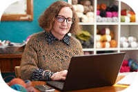 Retail worker in knit sweater types on laptop at desk surrounded by colorful yarn on shelves