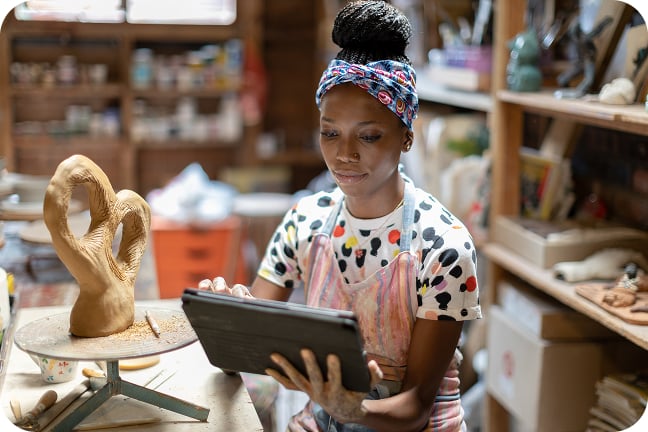 Small-business owner in pottery studio sits at table while looking at handheld tablet