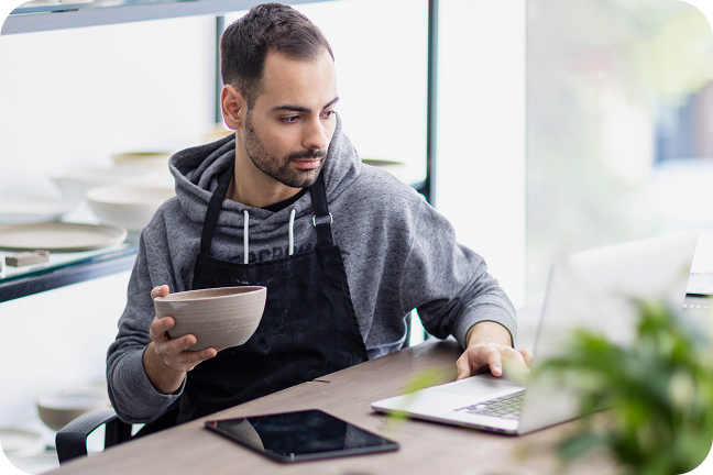 Retail worker in black apron holds ceramic bowl while working at wooden table with laptop and tablet