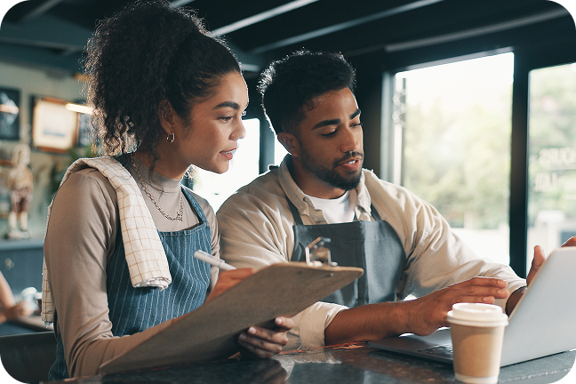 Two retail workers in aprons work together with laptop and clipboard inside small business