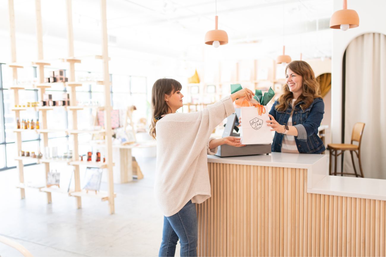 Retail store employee handing branded shopping bag across light-wood checkout counter to customer