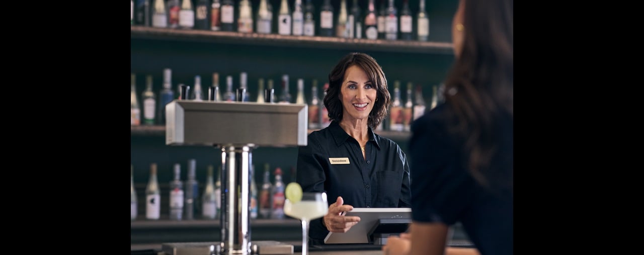 Smiling restaurant worker uses point of sale system while standing behind bar in server area